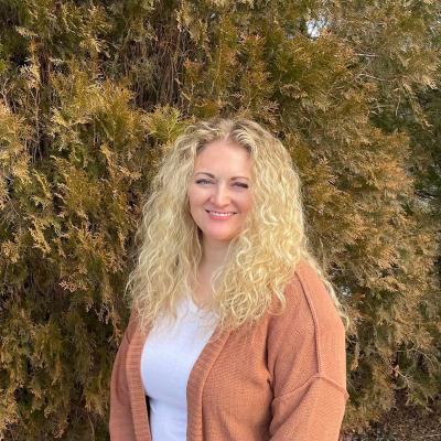 Photo of Dr. McKenna with blonde hair, wearing a white shirt and brown cardigan, with trees behind her