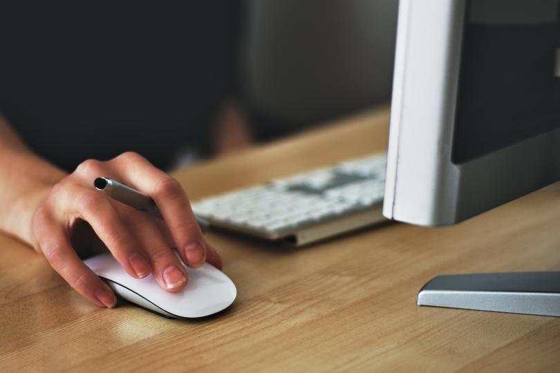 hand on mouse device holding pen on desk in front of desktop computer